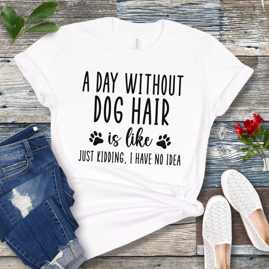 White dog lover t-shirt with the text 'A DAY WITHOUT DOG HAIR IS LIKE, JUST KIDDING, I HAVE NO IDEA' printed on it, displayed on a wooden surface with a pair of blue jeans and white shoes in the background.