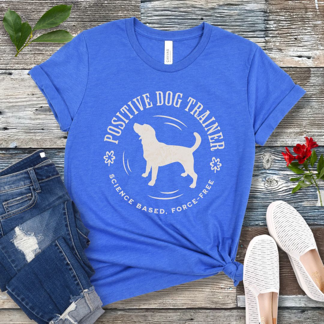 Royal blue t-shirt with 'Positive Dog Trainer, Science Based, Force Free' text and a silhouette of a dog in a circular design, displayed flat with a pair of blue jeans and white shoes on a wooden surface.