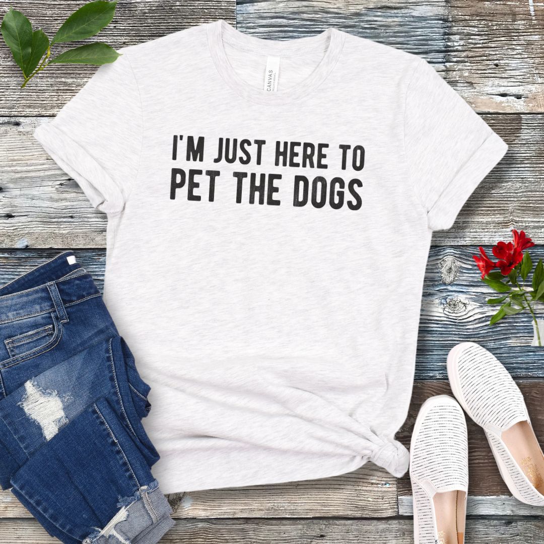 Light grey T-shirt with the text 'I'M JUST HERE TO PET THE DOGS' printed in white, displayed flat with a pair of blue jeans and white shoes.
