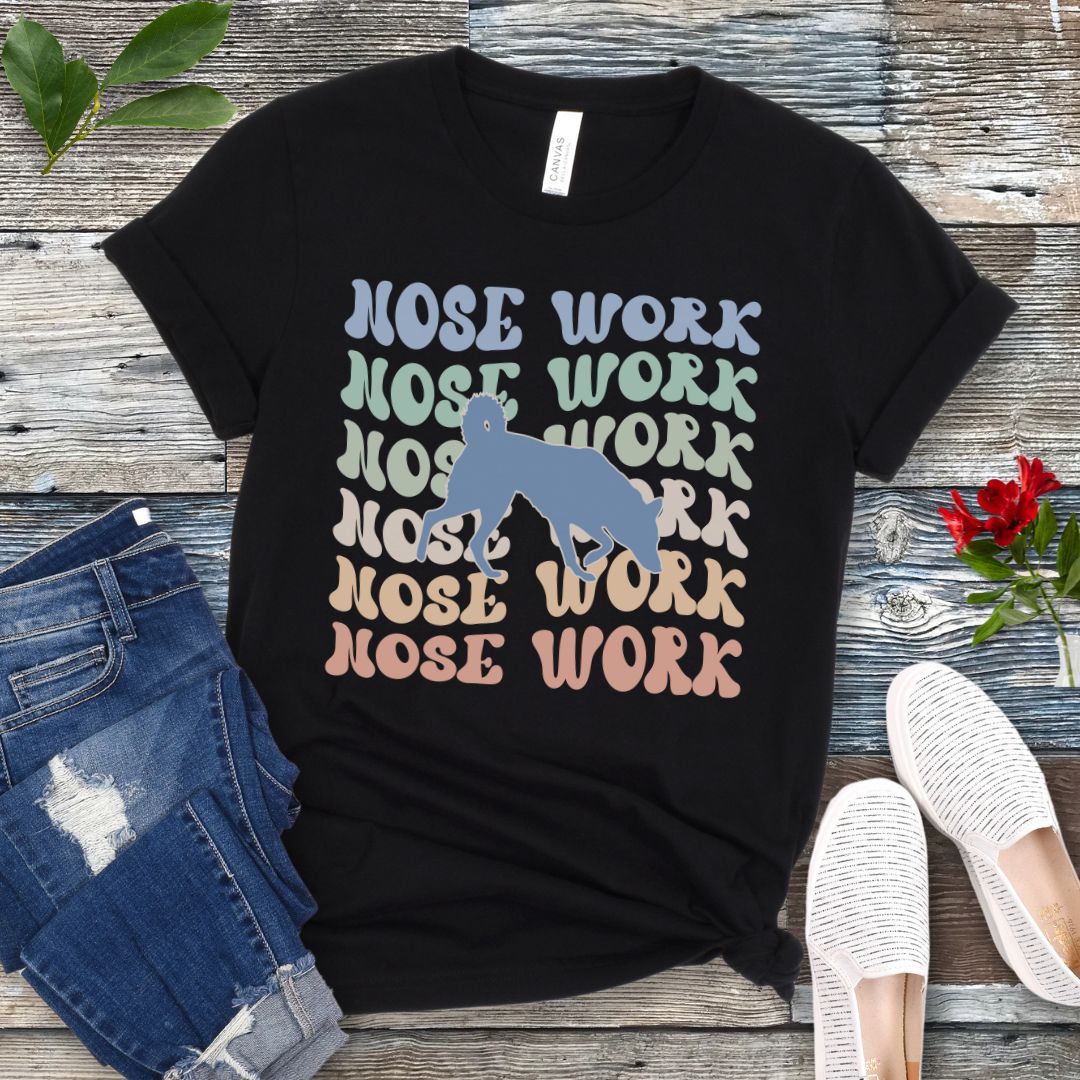 Black dog sports t-shirt with wavy multicolored 'NOSE WORK' text in a retro-style font and a custom dog breed silhouette, displayed flat with a pair of blue jeans and white shoes.