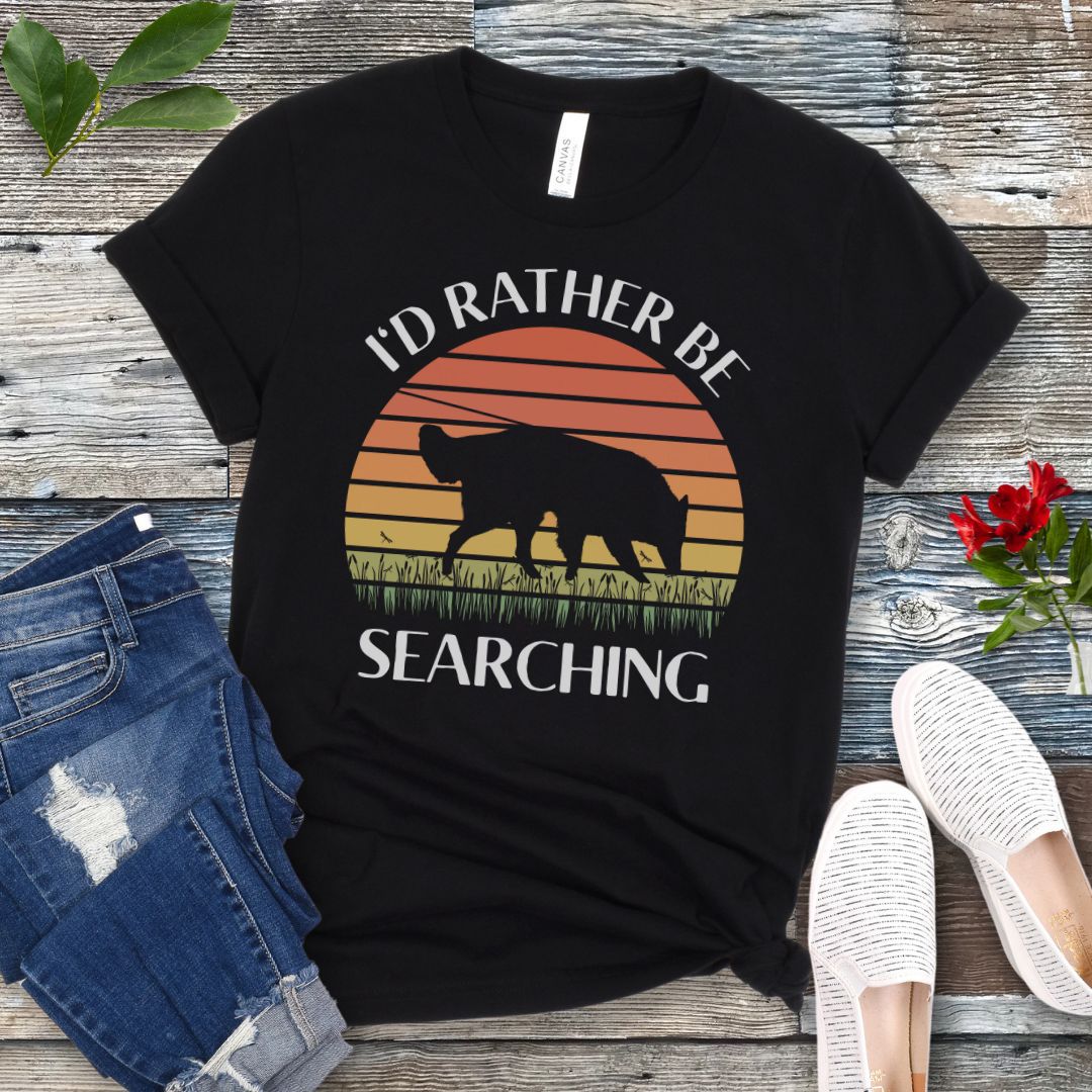 Black nosework t-shirt with custom breed sniffing dog graphic and 'I'd Rather Be Searching' text on a wooden surface with jeans and shoes.