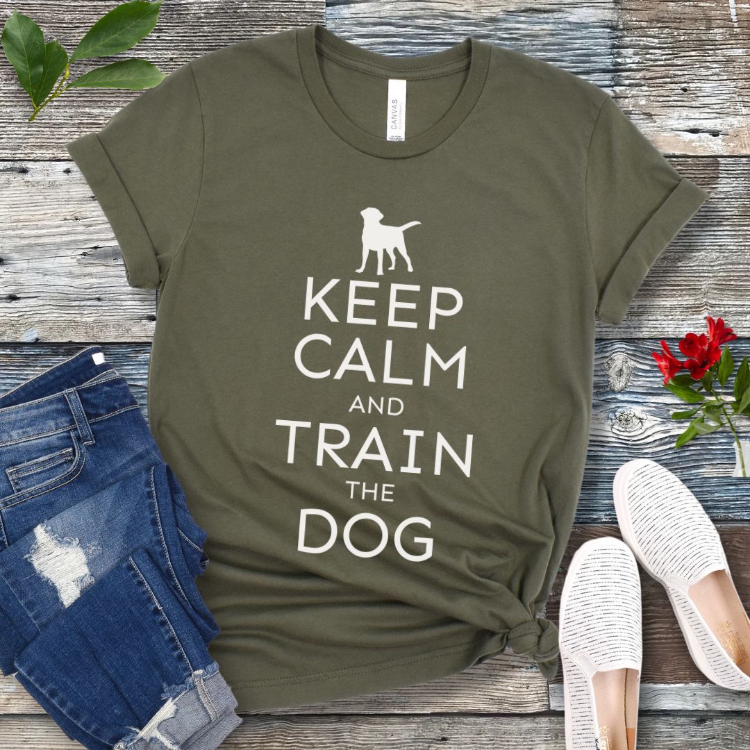 A military green t-shirt with white text and dog graphic and text that says 'KEEP CALM AND TRAIN THE DOG', displayed on a wooden background with a pair of blue jeans and white shoes.