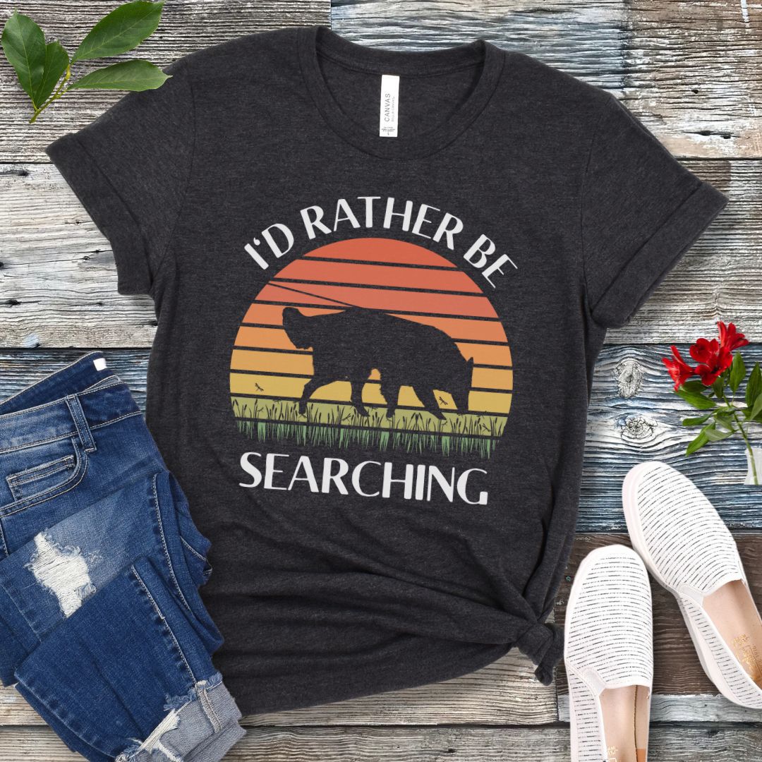Dark grey scentwork t-shirt with custom breed sniffing dog graphic and 'I'd Rather Be Searching' text on a wooden surface with jeans and shoes.