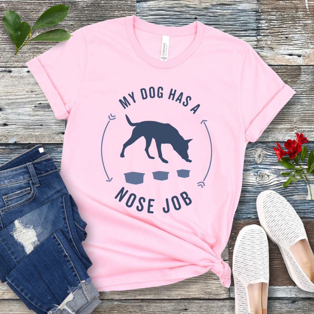 Pink canine scentwork t-shirt with graphic of a searching dog with text 'MY DOG HAS A NOSE JOB' on the front, presented on a wooden surface with denim jeans and white shoes in the background.