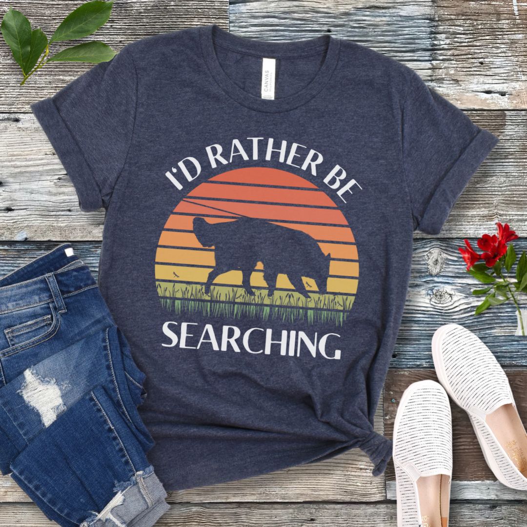 Heather navy blue scent detection t-shirt with custom breed sniffing dog graphic and 'I'd Rather Be Searching' text on a wooden surface with jeans and shoes