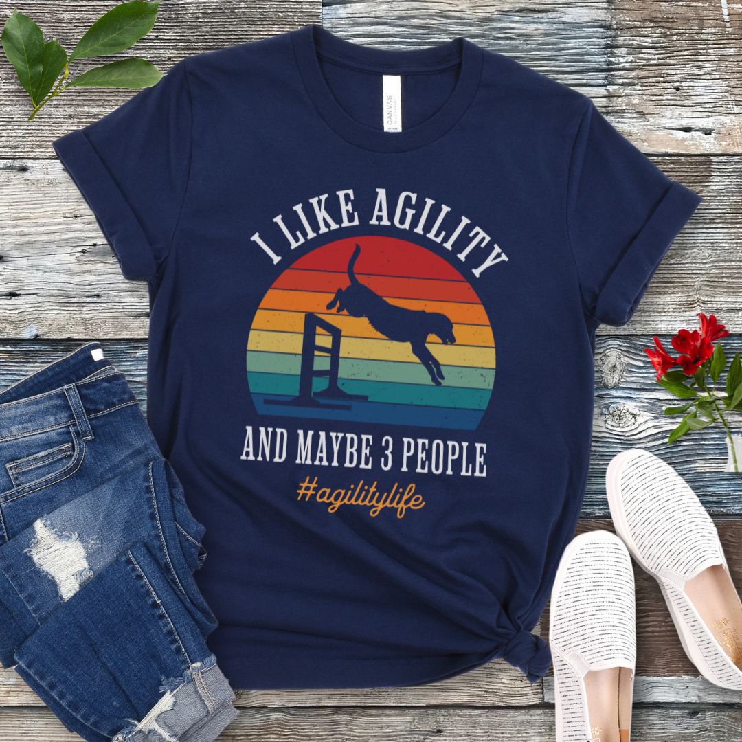 Navy blue dog sports t-shirt with jumping dog image and the text 'I Like Agility and Maybe 3 People' on a wooden surface with jeans and shoes.