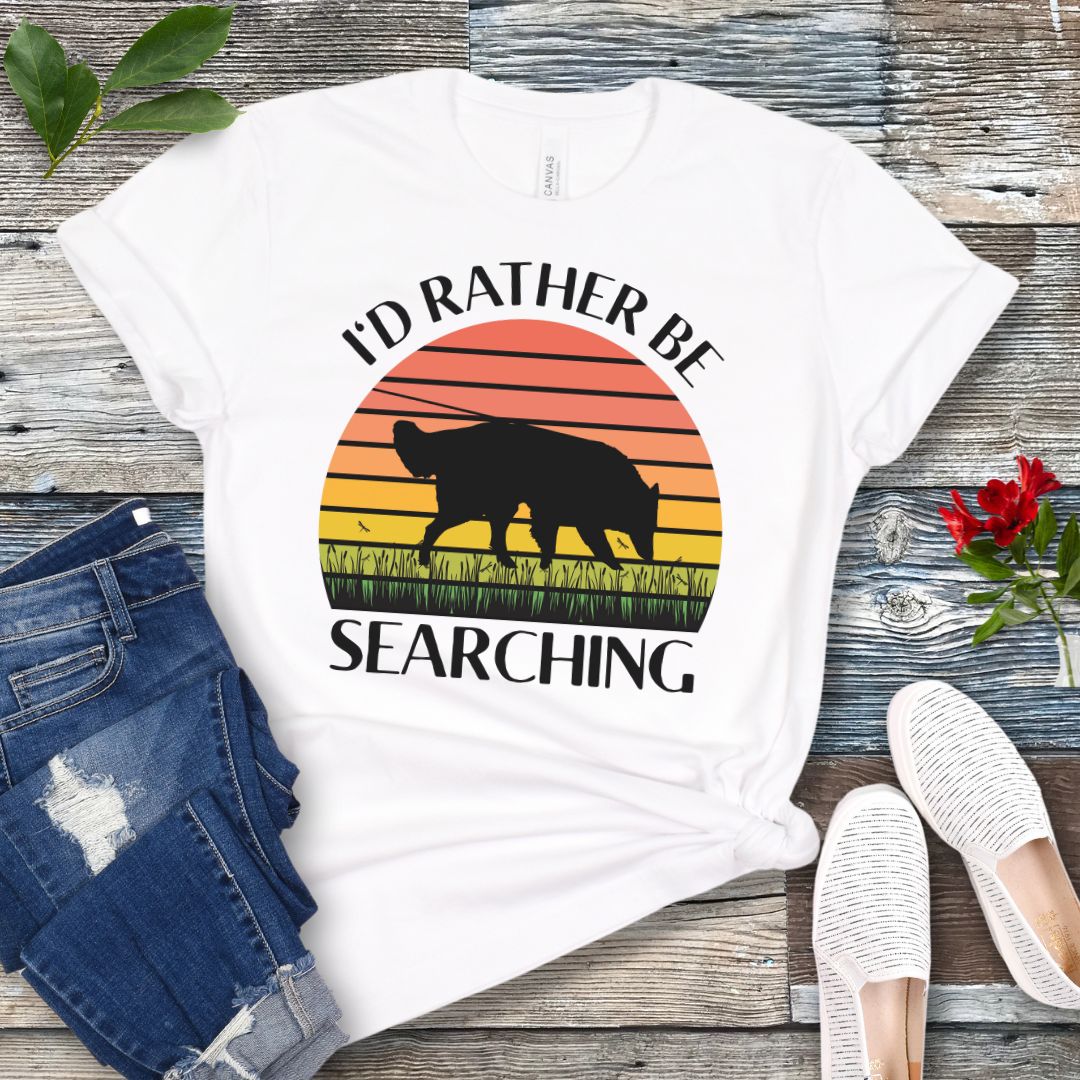 White nosework t-shirt with custom breed sniffing dog graphic and 'I'd Rather Be Searching' text on a wooden surface with jeans and shoes.