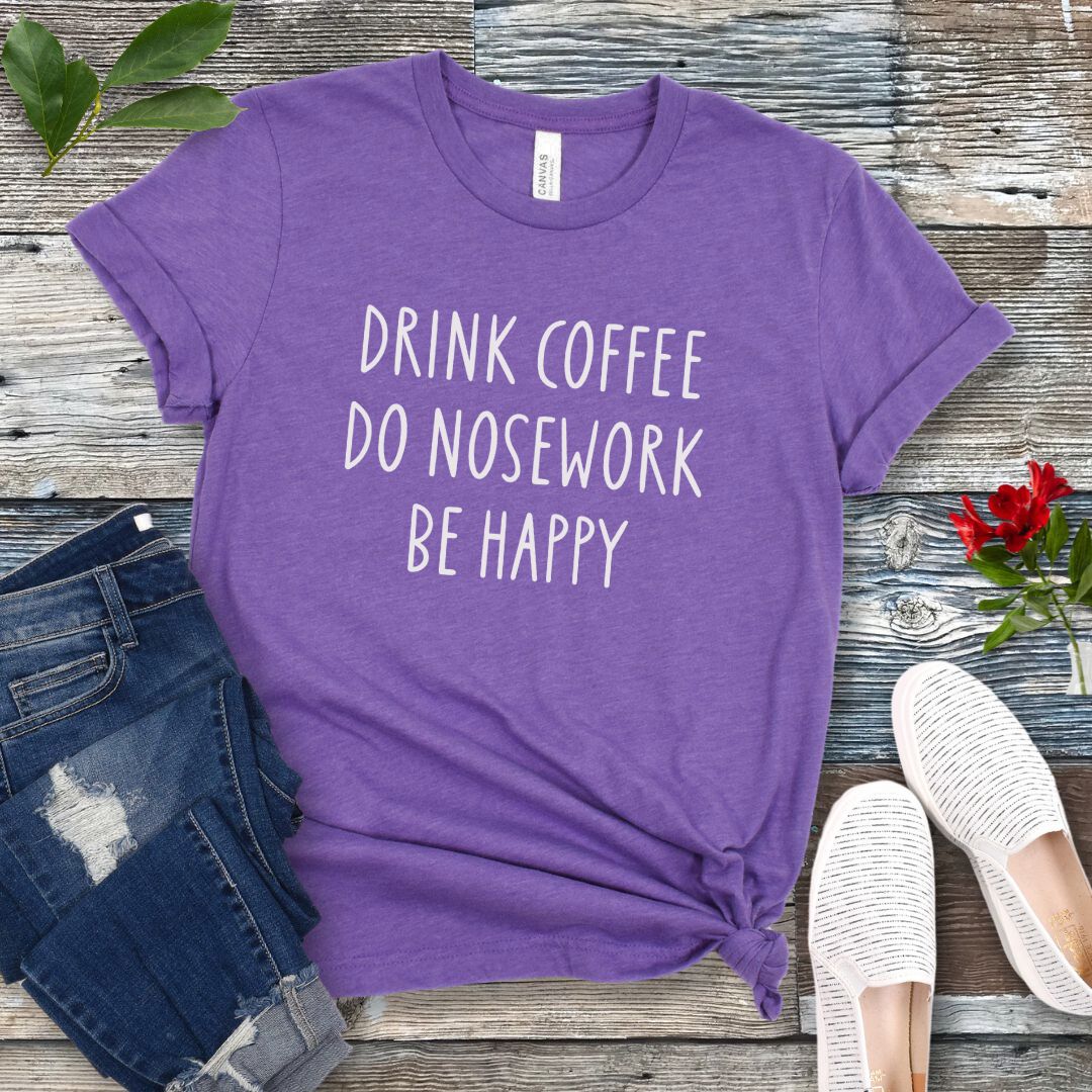 Purple dog sports t-shirt with text 'Drink coffee, do nosework, be happy' on a wooden surface with jeans and shoes.