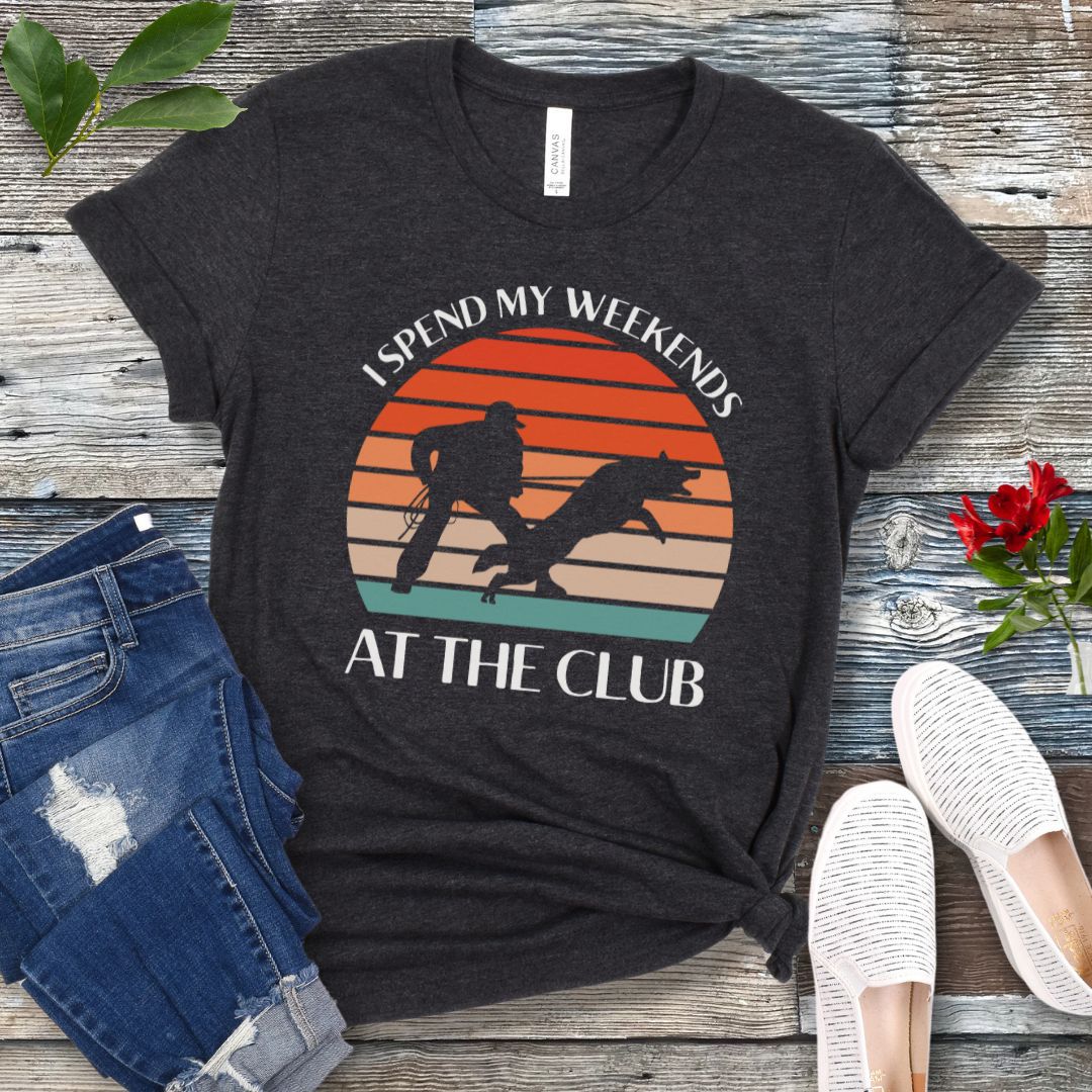 Dark grey Schutzhund t-shirt with graphic of a dog and handler doing bitework and text 'I SPEND MY WEEKENDS AT THE CLUB' on a wooden background, accompanied by denim jeans and white shoes.