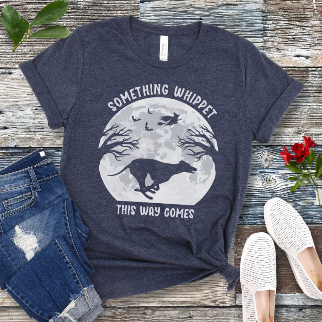 A heather navy t-shirt with a Halloween graphic of a full moon, flying witch, bats and running whippet, and the text 'something whippet this way comes', displayed flat with a pair of jeans and white shoes.
