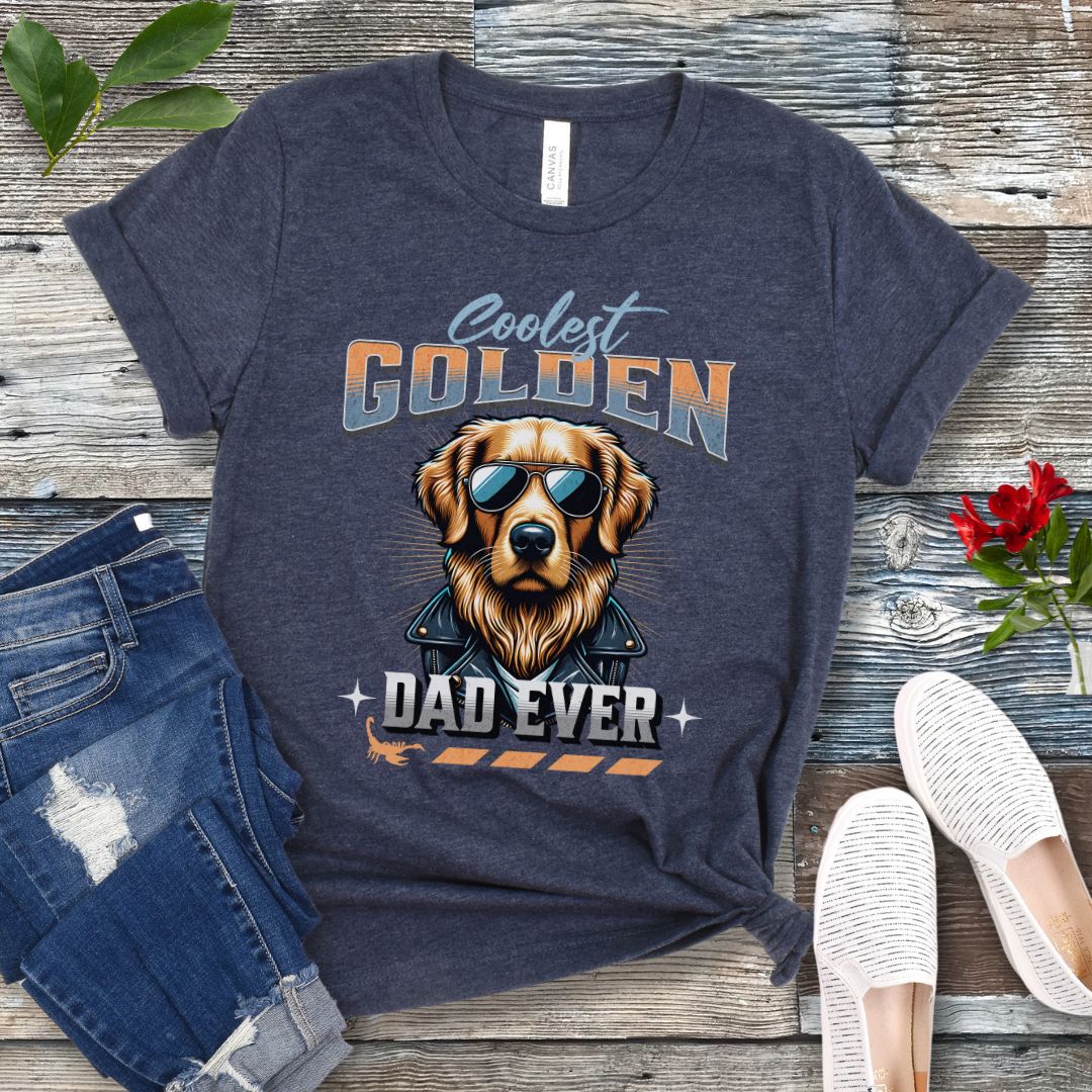 A heather navy blue t-shirt with a graphic print of a Golden Retriever dog wearing sunglasses and leather jacket and the text 'Coolest Golden Dad Ever', presented on a wooden surface with a pair of blue jeans and white shoes.