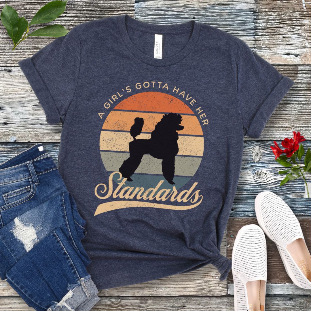 Navy blue heather t-shirt with a graphic of a poodle and the text 'A Girl's Gotta Have Her Standards', presented on a wooden surface with denim jeans and white shoes in the background.