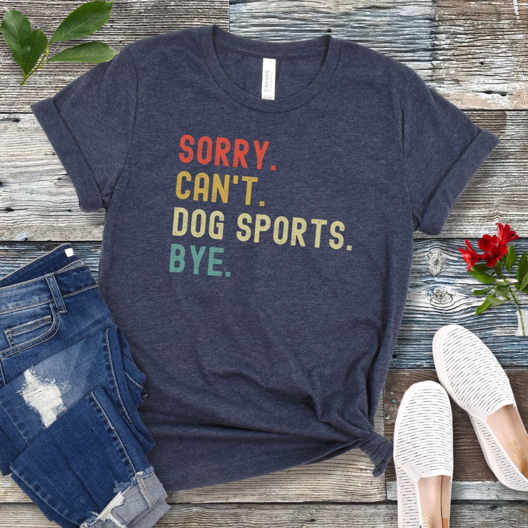 Heather navy t-shirt with the text 'SORRY, CAN'T, DOG SPORTS, BYE' printed in colorful letters on a wooden background, accompanied by casual clothing and shoes.