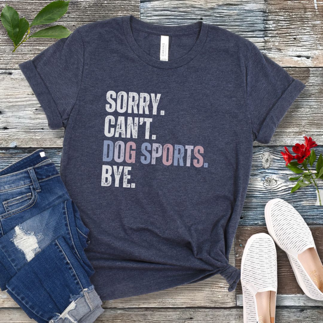 Heather navy t-shirt with the text 'SORRY. CAN'T. DOG SPORTS. BYE.' in blocky, distressed lettering, presented on a wooden surface with jeans and casual shoes.