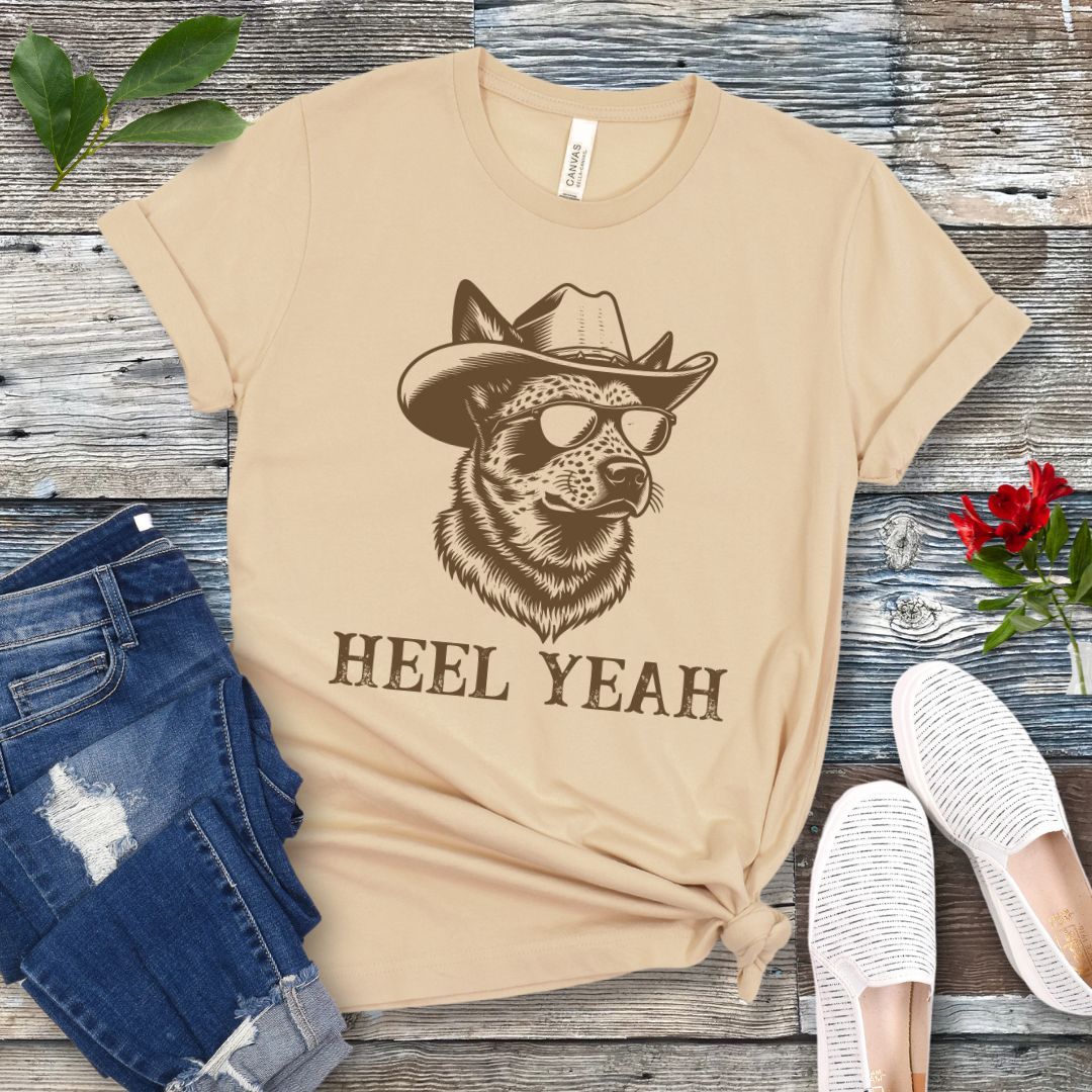 Cream t-shirt with a graphic of a Cattle dog wearing sunglasses and a cowboy hat, text 'HEEL YEAH', displayed on a wooden surface with jeans and shoes.