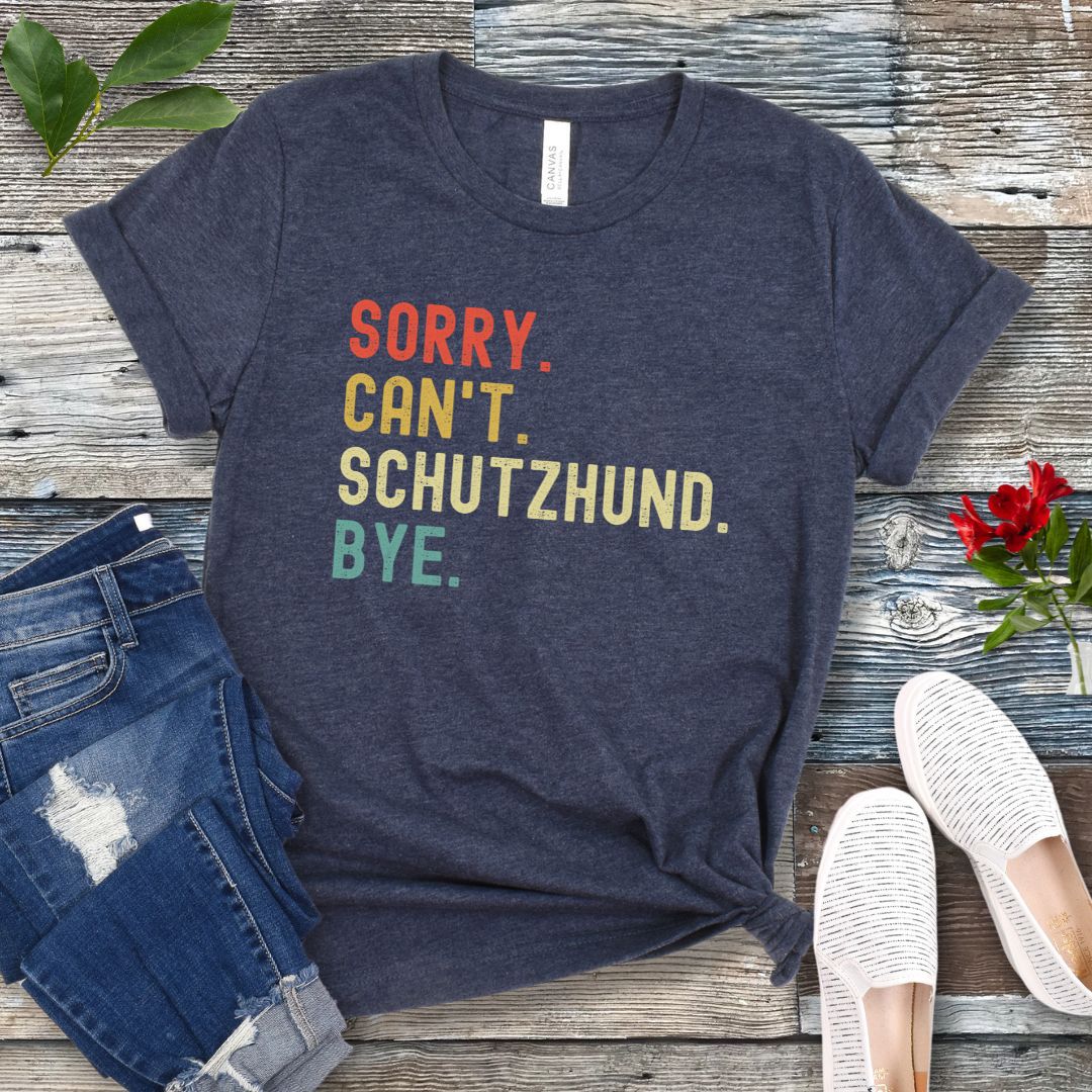 Heather navy t-shirt with colorful text 'SORRY. CAN'T. SCHUTZHUND.BYE.' presented on a wooden surface with jeans and casual shoes.