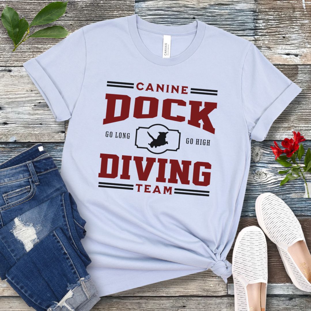 A llght blue t-shirt with 'CANINE DOCK DIVING TEAM' text and a graphic of a dock jumping dog, placed on a wooden surface with jeans and white shoes in the background.