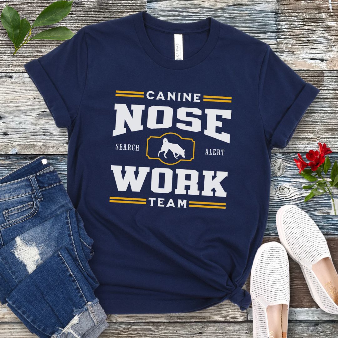 A navy blue dog sports t-shirt with 'CANINE NOSE WORK TEAM' printed on the front and custom dog breed silhouette, laid out flat with a pair of blue jeans and white shoes on a wooden surface.