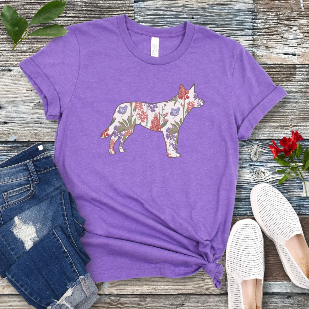 A purple t-shirt with a floral-filled silhouette of a cattle dog, accompanied by a pair of rolled-up blue jeans and a pair of white shoes on a wooden surface.