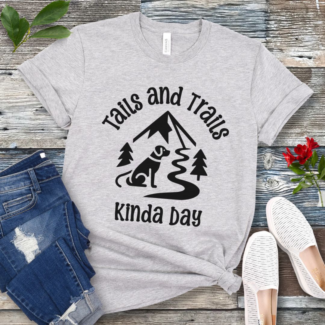 A grey t-shirt with stylized graphic of dog, trees and mountain, and the text 'Tails and Trails Kinda Day', presented on a wooden surface with jeans and casual shoes.