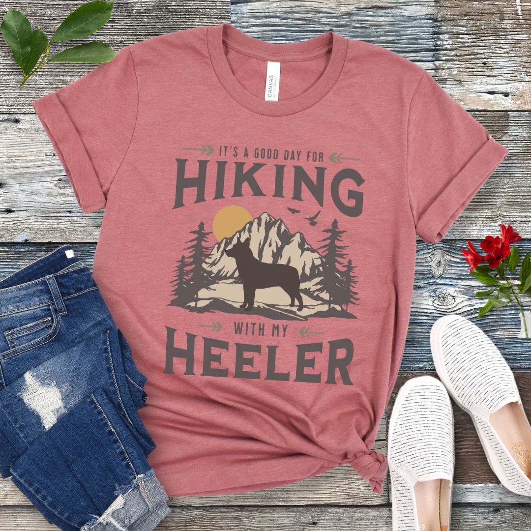 Mauve dog hiking t-shirt with a graphic print of a dog and mountains, and text that reads 'HIKING WITH MY HEELER'. The shirt is presented on a wooden surface with a pair of blue jeans and white shoes in the background.