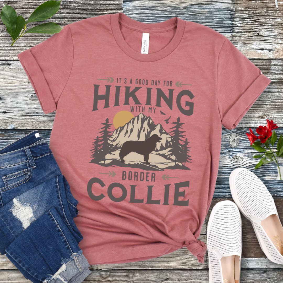 Mauve unisex short sleeve t-shirt with a graphic print of a Border Collie in a mountain scene and text 'It's a good day for hiking with my BORDER COLLIE', displayed on a wooden surface with jeans and white shoes in the background.