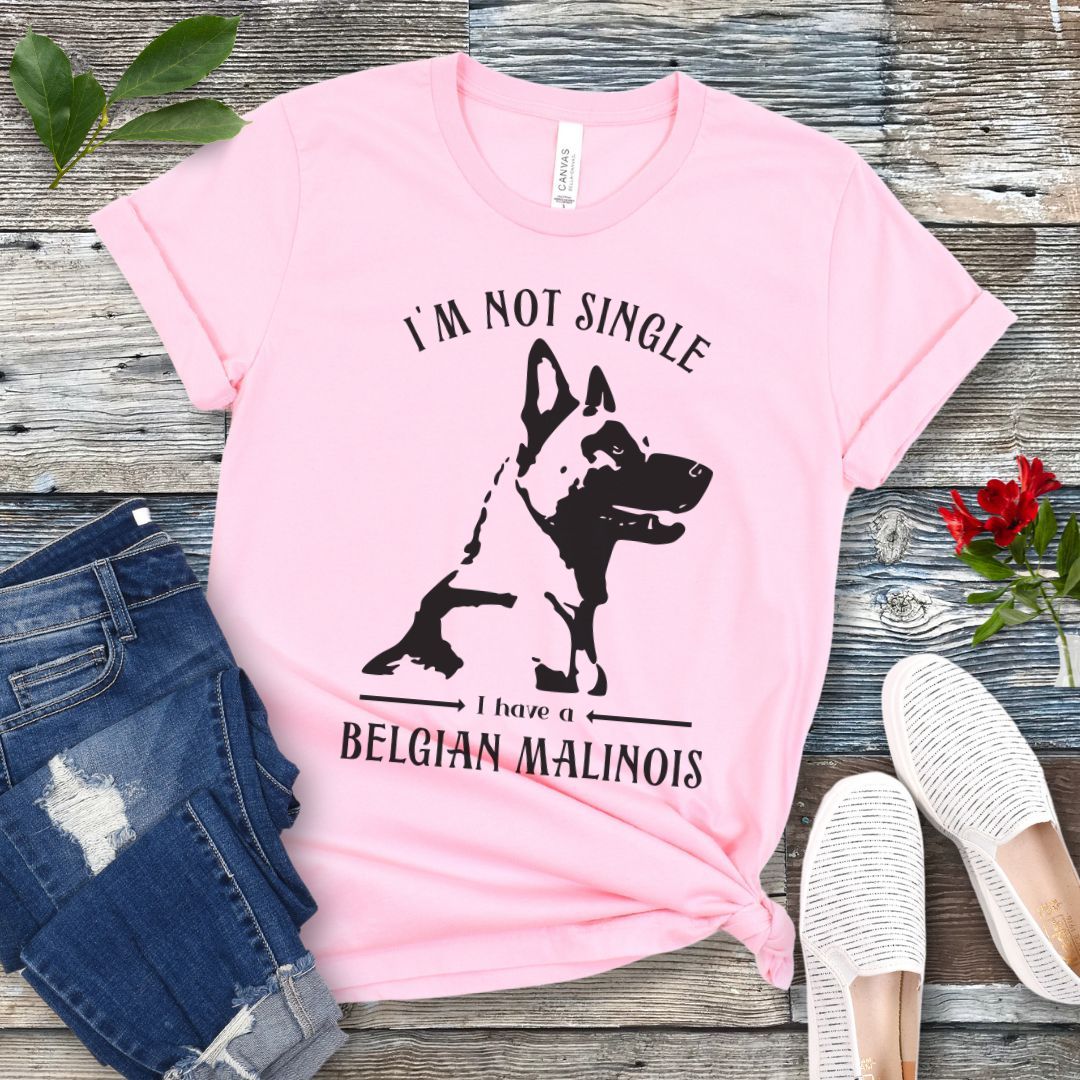 Pink t-shirt with "I'm Not Single I have a Belgian Malinois" text and silhouette of a Belgian Malinois dog printed on it, displayed flat with a pair of jeans and white shoes on a wooden surface.