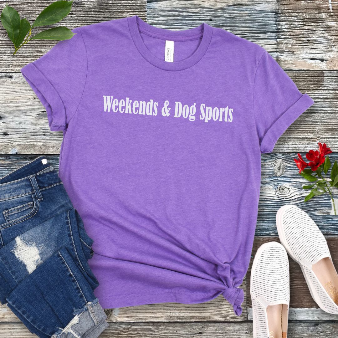 A purple Bella Canvas 3001 unisex short sleeve t-shirt with the text 'Weekends & Dog Sports' printed on the front, displayed on a wooden surface with a pair of blue jeans and white shoes in the background.