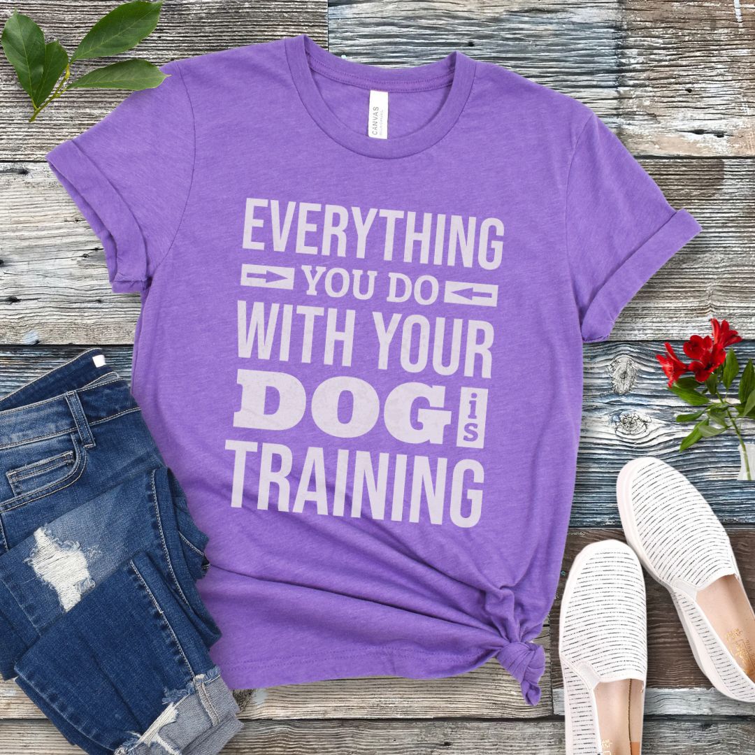 Purple t-shirt with the text 'EVERYTHING YOU DO WITH YOUR DOG IS TRAINING' text design, displayed flat with a pair of blue jeans and white shoes.