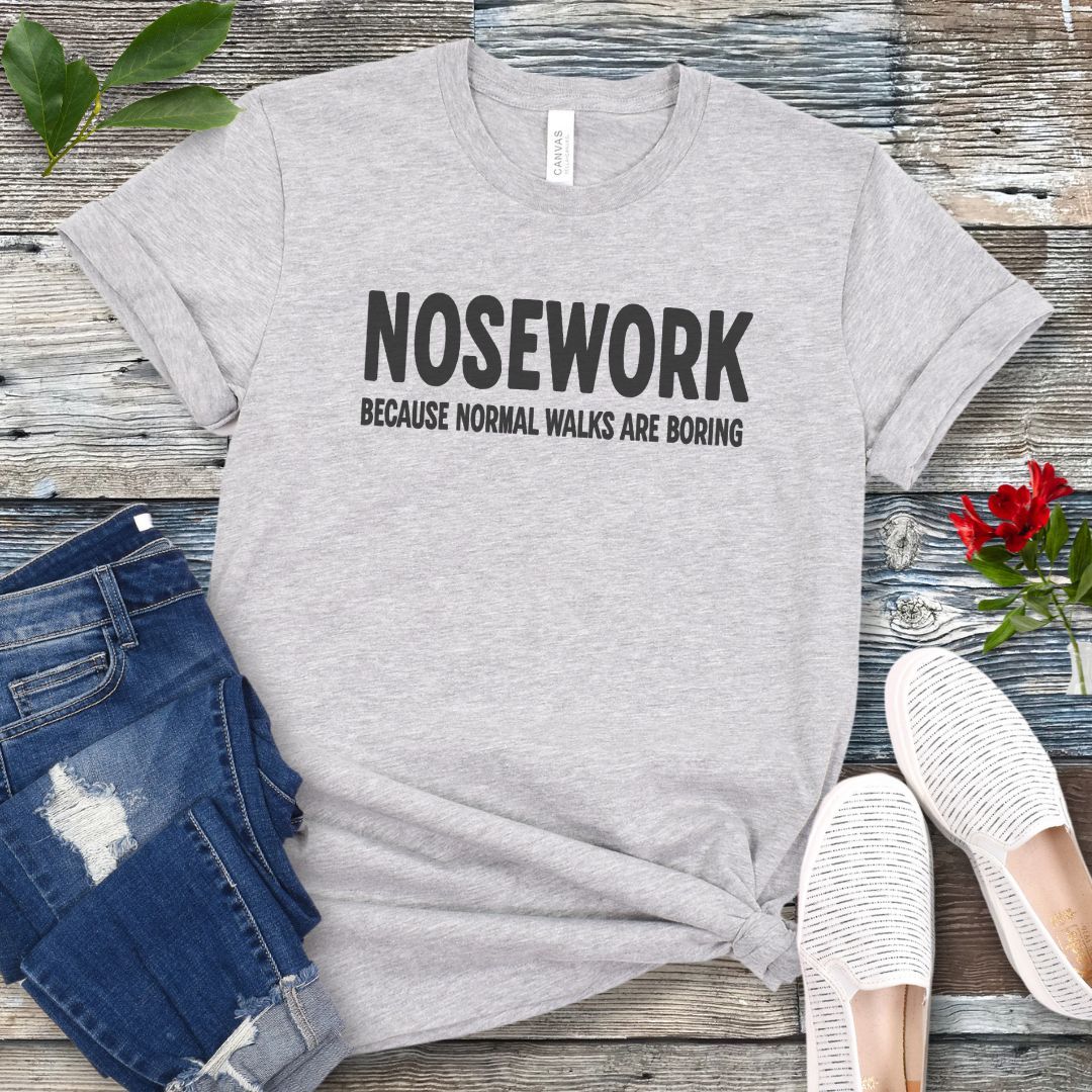 Light grey K9 Nose Work t-shirt with 'NOSEWORK BECAUSE NORMAL WALKS ARE BORING' text on a wooden surface with jeans and shoes.
