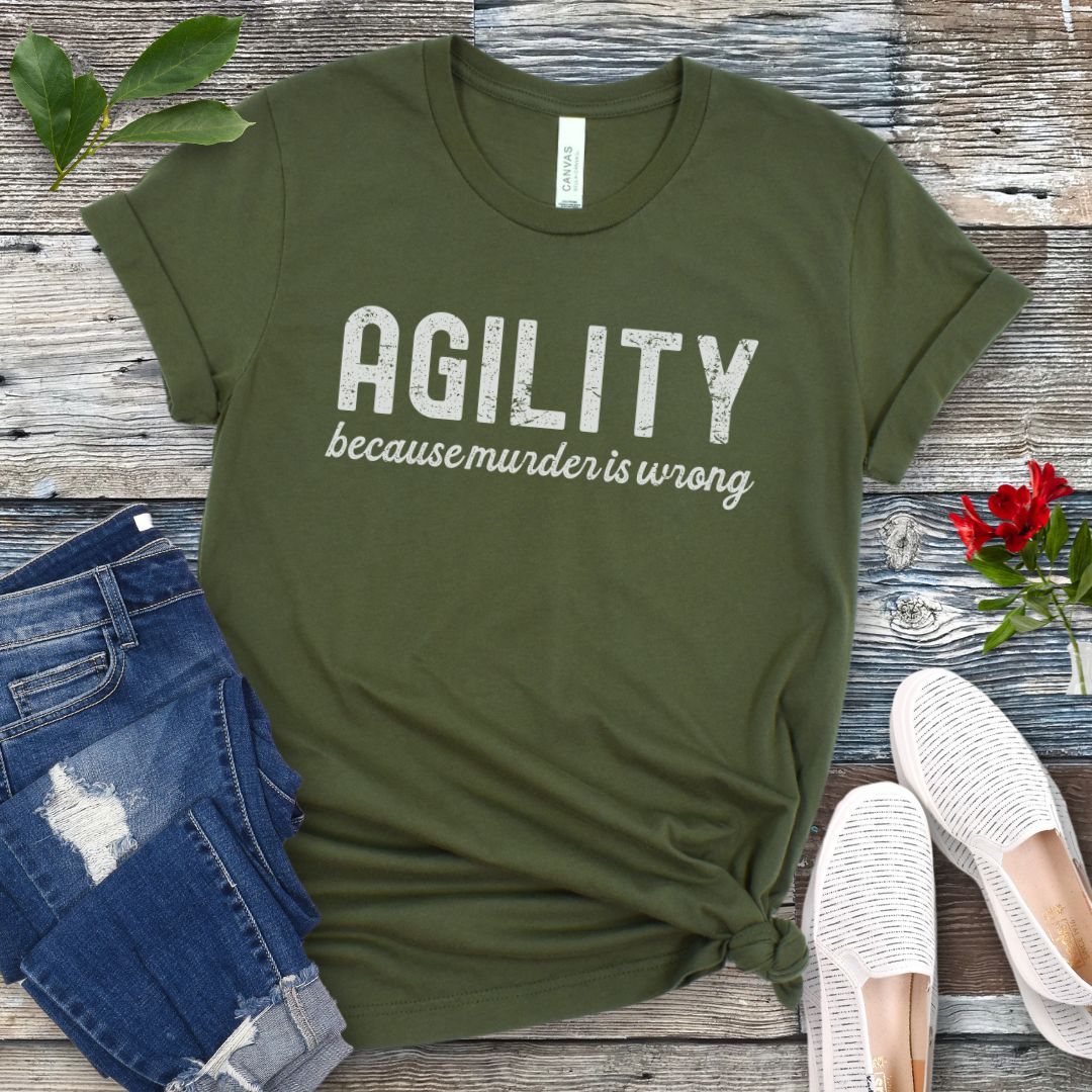 Military green t-shirt with 'AGILITY because murder is wrong' text on a wooden surface with jeans and shoes.