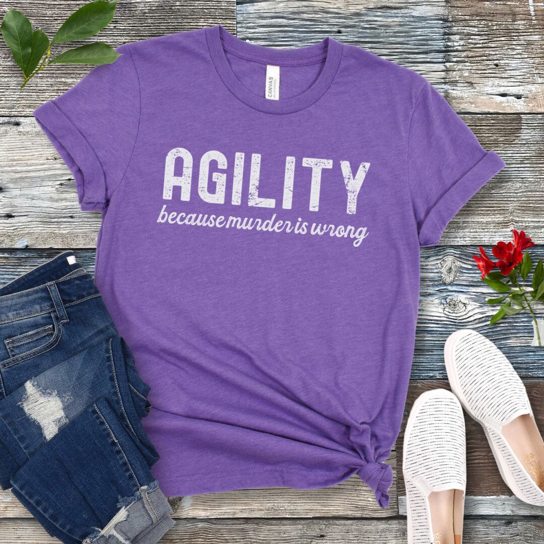 Purple t-shirt with 'AGILITY because murder is wrong' text on a wooden surface with jeans and shoes.