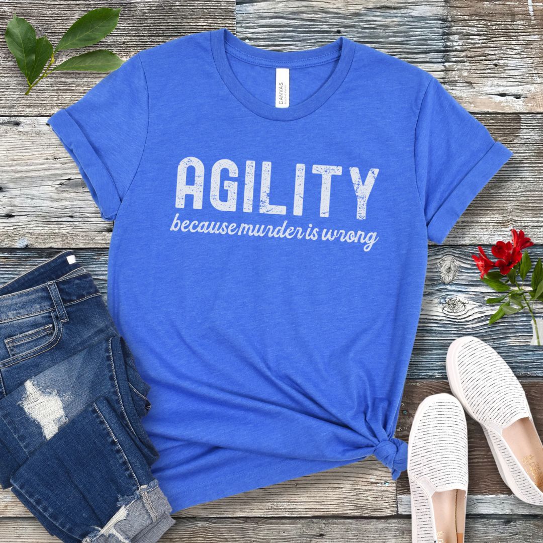 Blue t-shirt with 'AGILITY because murder is wrong' text on a wooden surface with jeans and shoes.