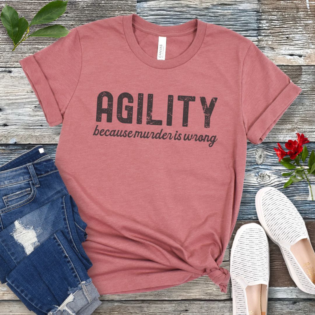 Mauve t-shirt with 'AGILITY because murder is wrong' text on a wooden surface with jeans and shoes.