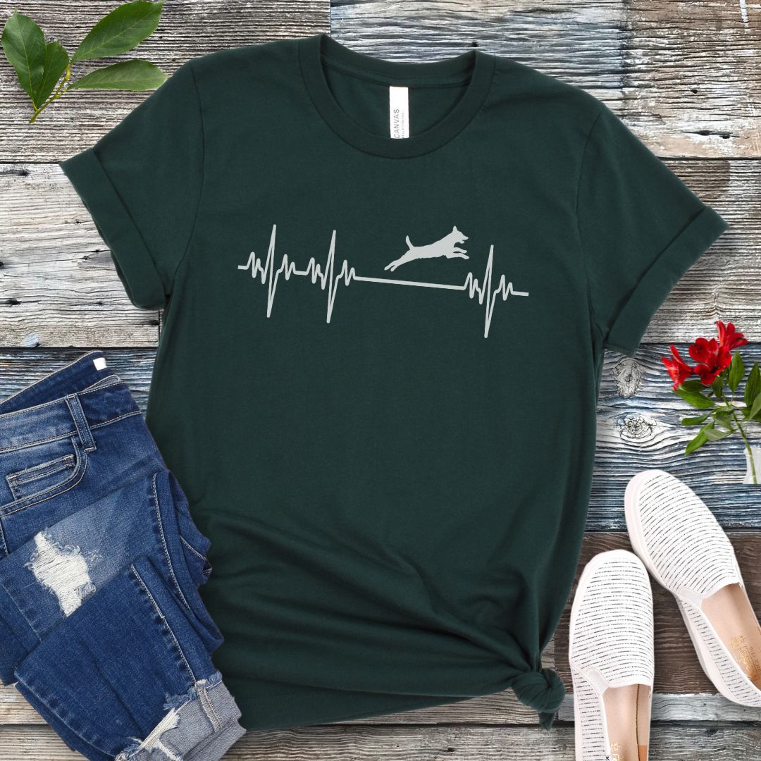 Dark green agility t-shirt with a heartbeat/EKG design with agility dog on a wooden surface with jeans and shoes.