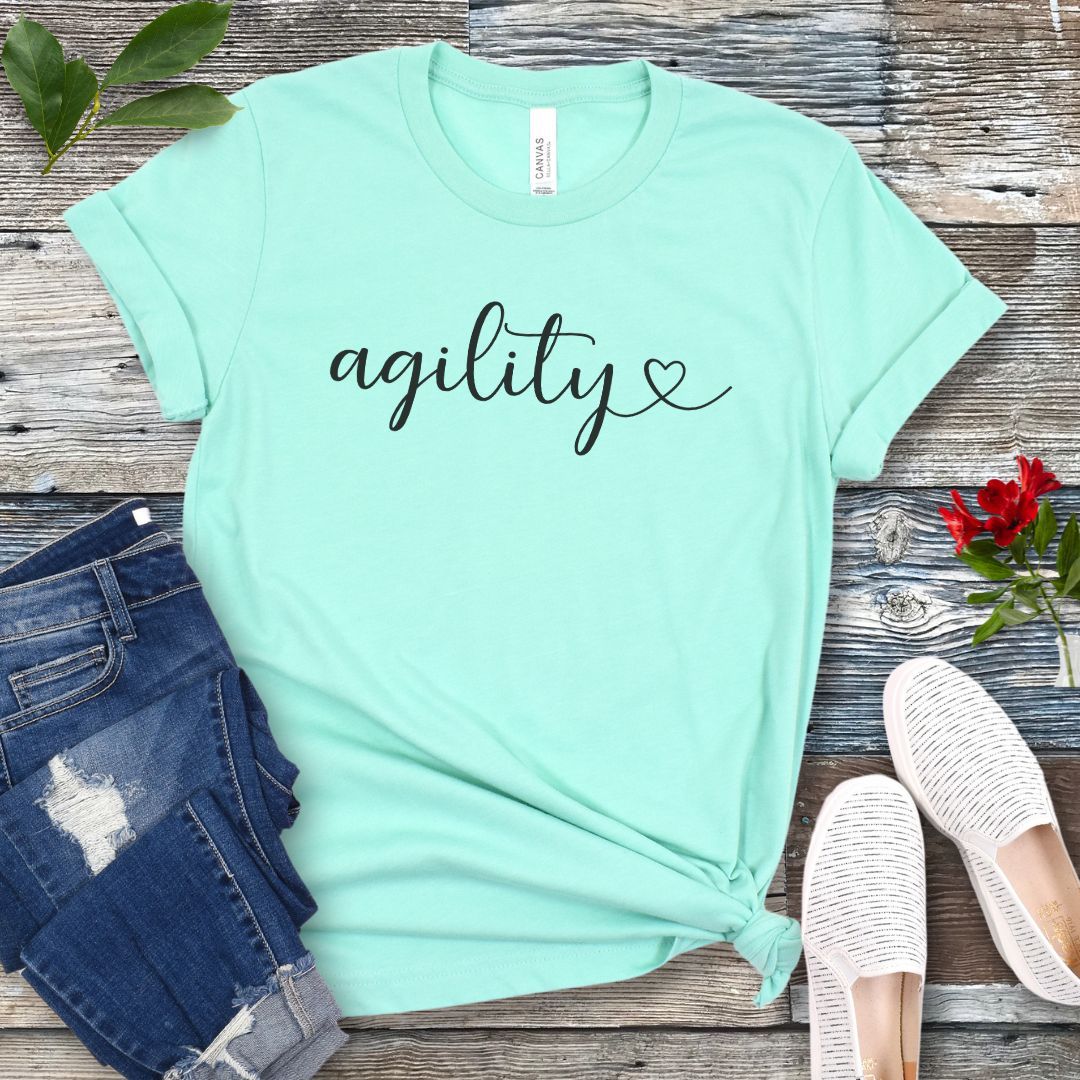 Mint green dog agility t-shirt with 'agility' in handwritten script on a wooden surface with jeans and shoes.