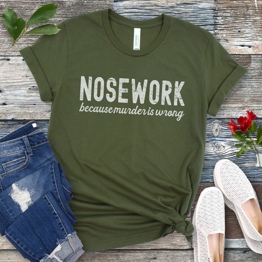 Military green dog sports t-shirt with 'Nosework because murder is wrong' text on a wooden surface with jeans and shoes.