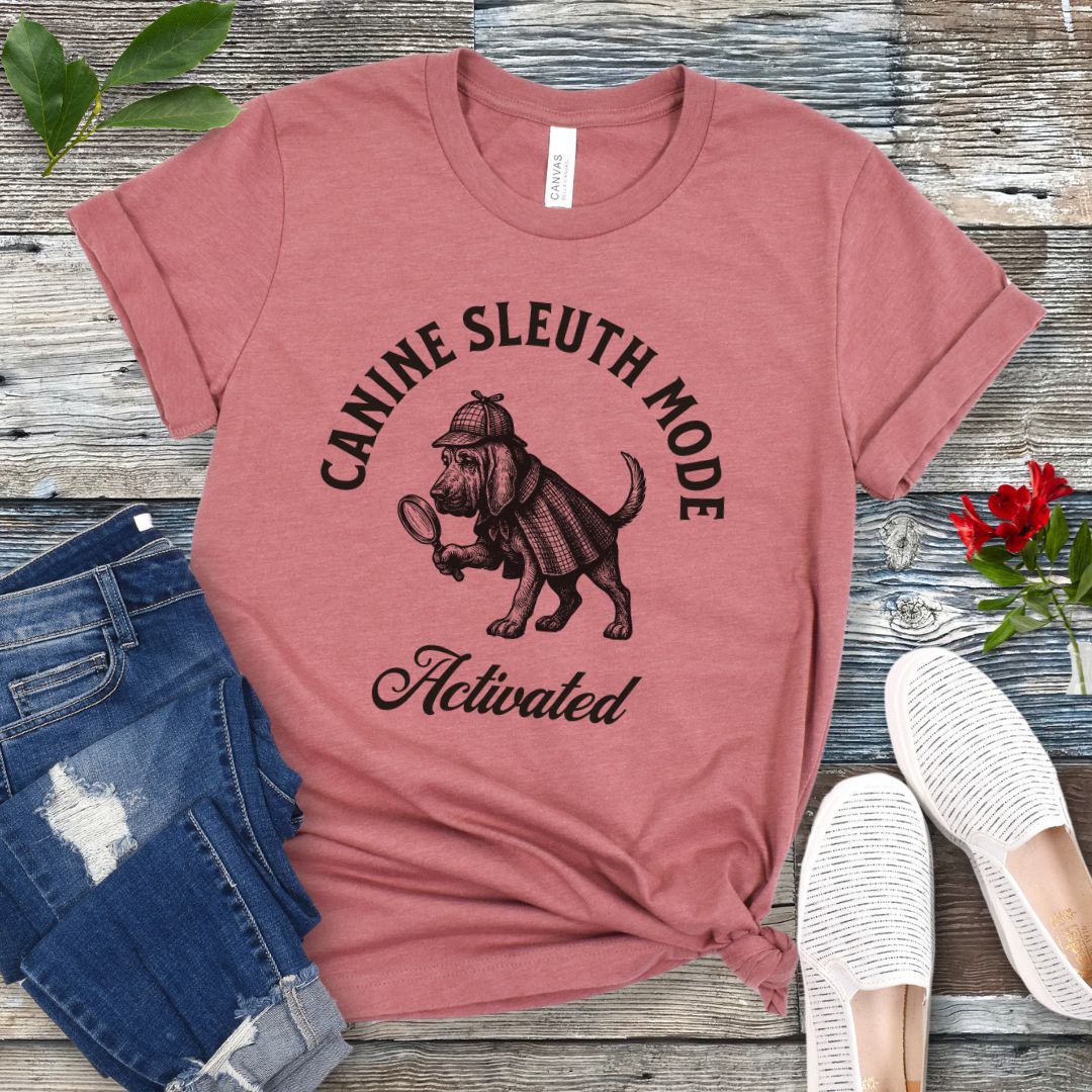 Mauve scent detection t-shirt with a vintage style graphic of a bloodhound in Sherlock Holmes' apparel, and 'Canine Sleuth Mode - Activated' text , presented on a wooden surface with jeans and shoes.