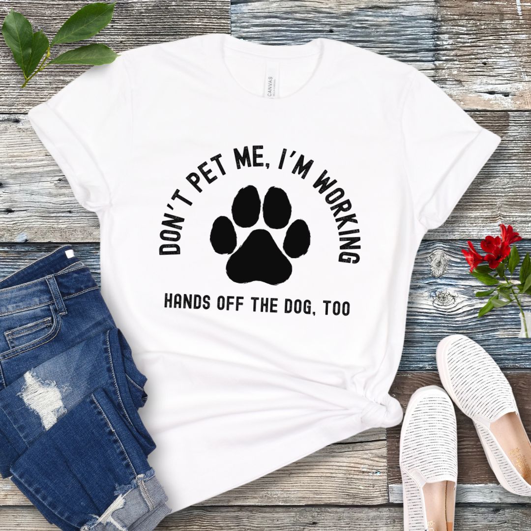 A white T-shirt with the text 'DON'T PET ME, I'M WORKING. HANDS OFF THE DOG, TOO' and a paw print design, displayed on a wooden surface with a pair of jeans and white shoes in the background.