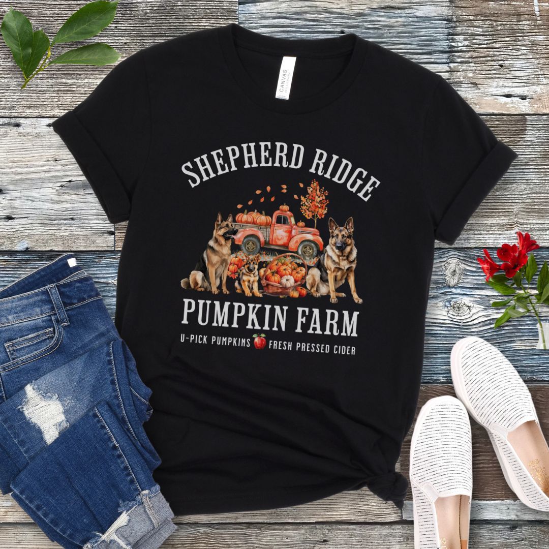 Black t-shirt with watercolor graphic of German Shepherds, a pumpkins truck and fall leaves, with the text 'Shepherd Ridge Pumpkin Farm', presented on a wooden surface with jeans and shoes.