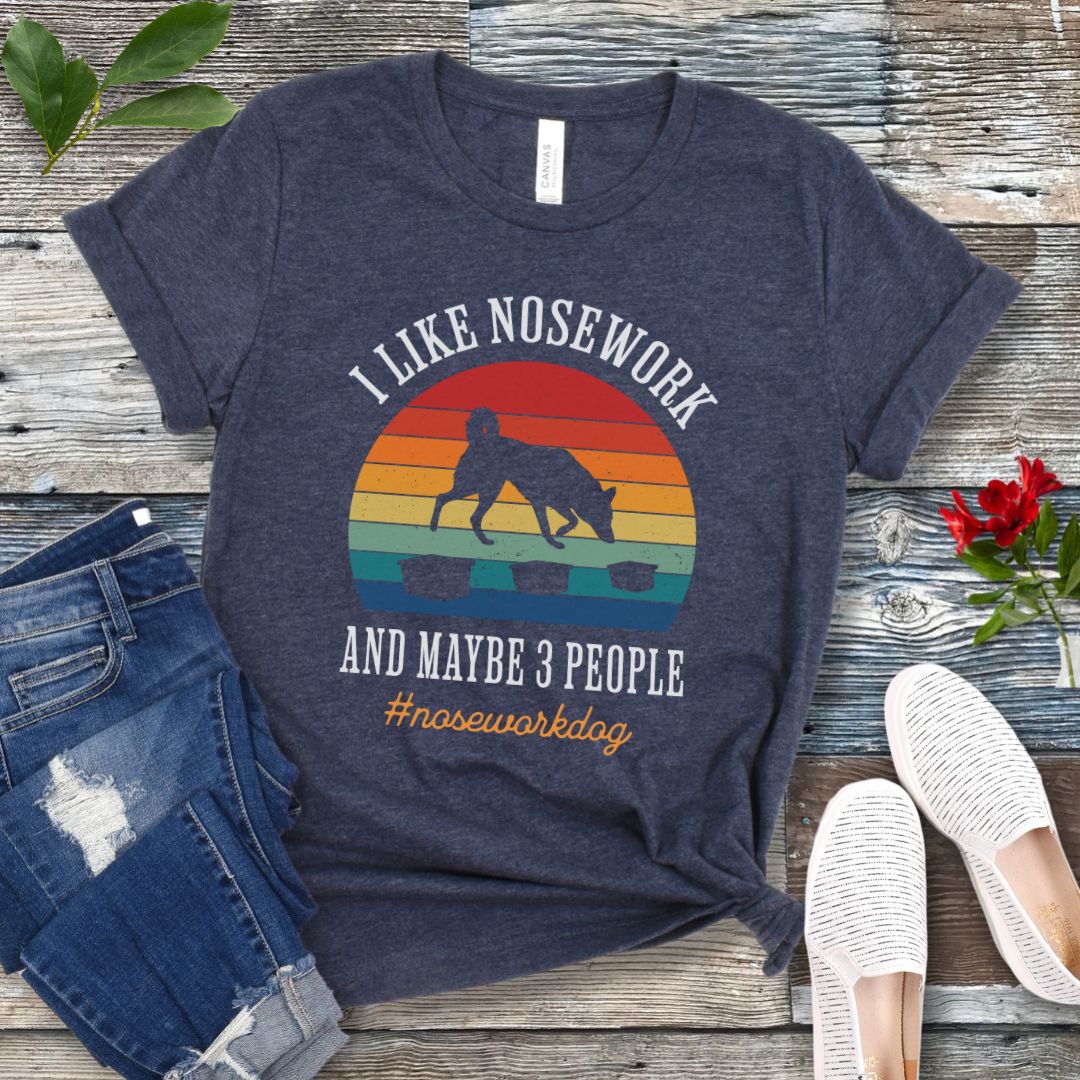 Heather navy blue nosework t-shirt with sniffing dog graphic and "I Like Nosework and Maybe 3 People" text on a wooden surface with jeans and shoes.