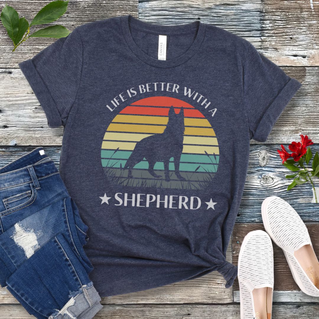 Heather navy t-shirt with colorful vintage sunset, German Shepherd graphic and the text 'Life is Better with a Shepherd', presented on a wooden surface with jeans and shoes.
