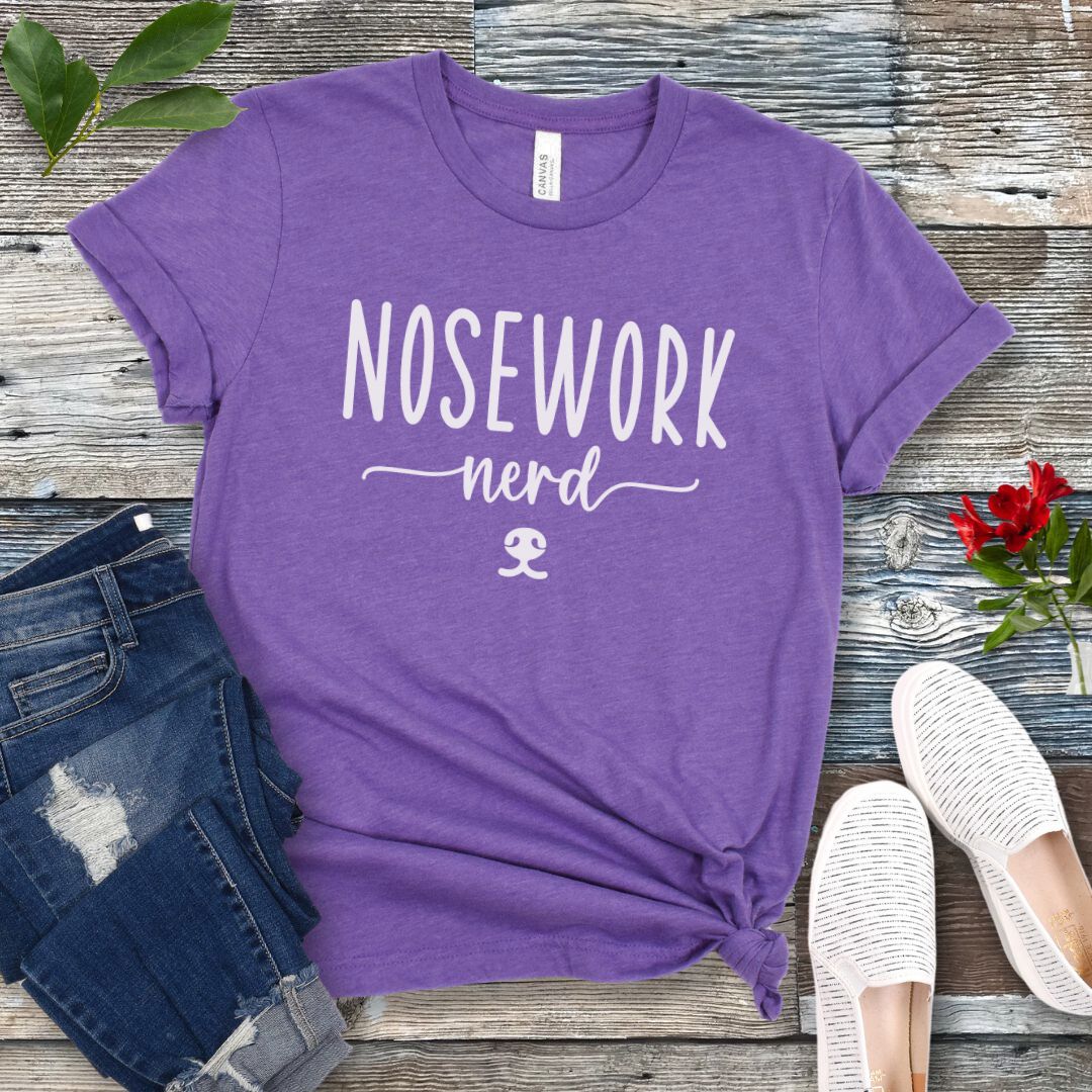 Purple dog sports t-shirt with 'Nosework nerd' text on a wooden surface with jeans and shoes.
