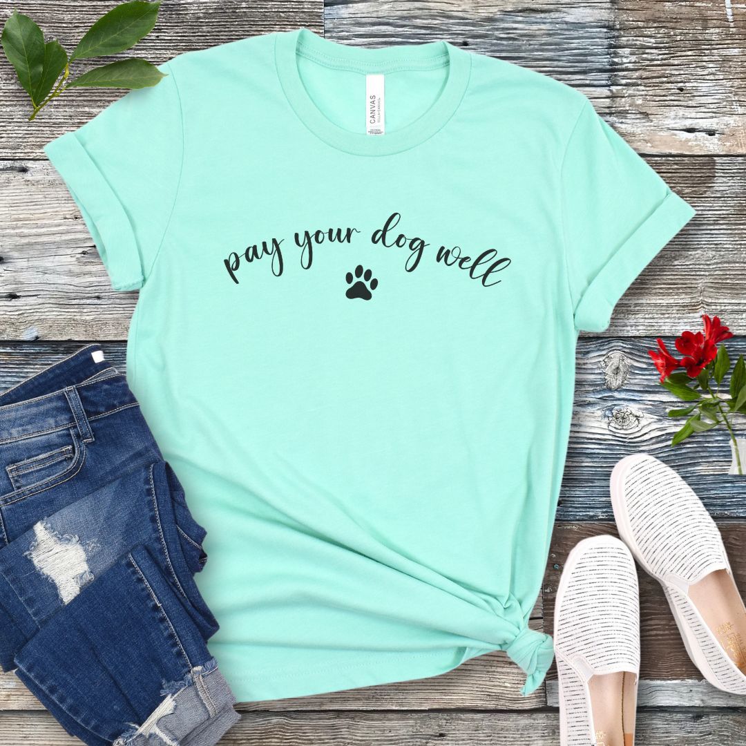 Mint green dog training t-shirt with 'pay your dog well' text and paw print on a wooden surface with jeans and shoes.