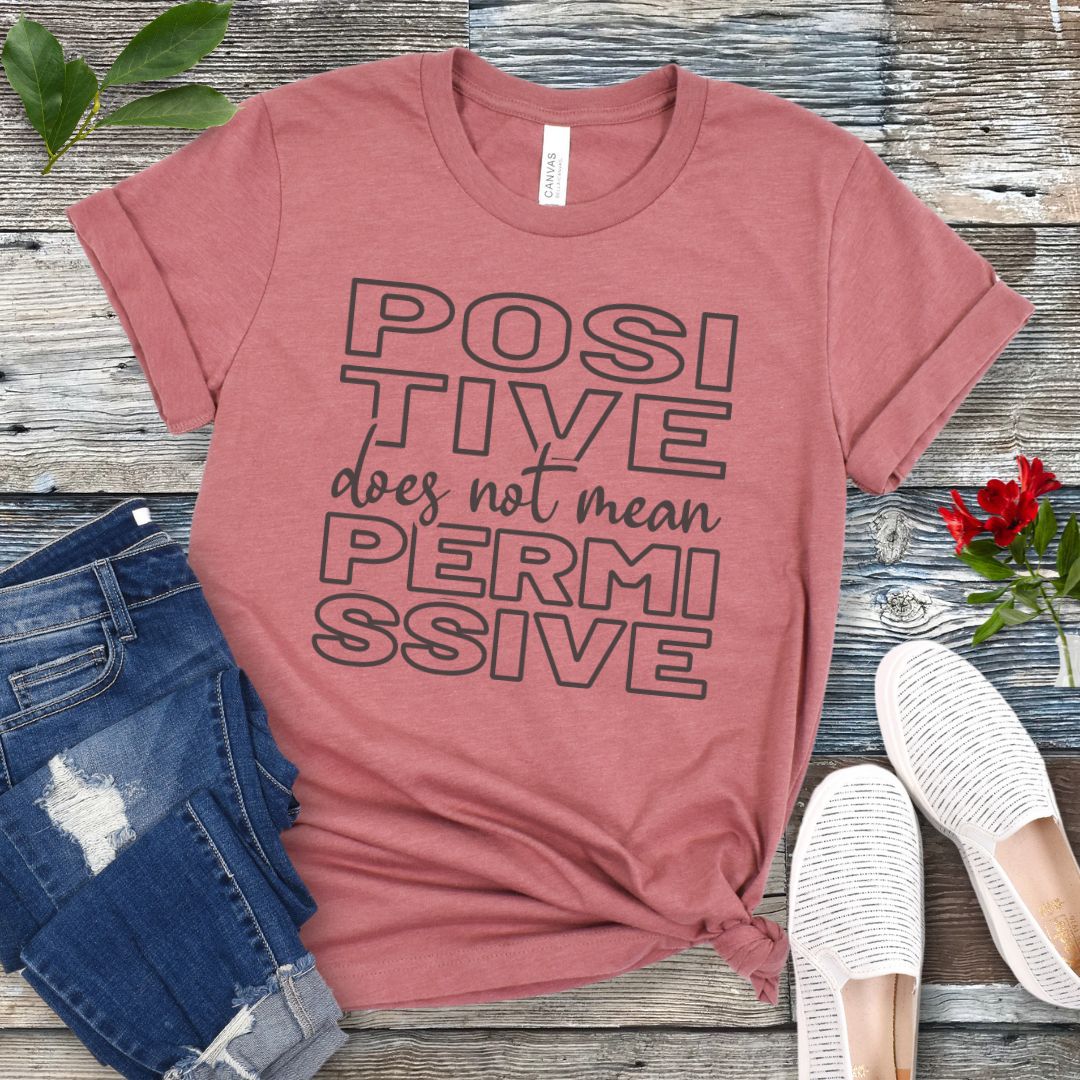 Mauve dog training t-shirt with 'POSITIVE does not mean PERMISSIVE' typography text on a wooden surface with jeans and shoes.