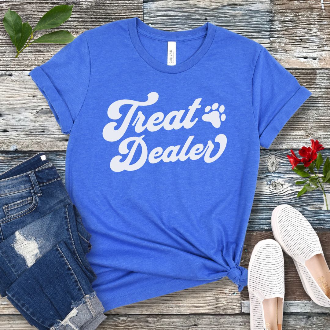 A blue T-shirt with the retro font with the text 'Treat Dealer' printed on the front, accompanied by a paw print. The shirt is displayed on a wooden background with a pair of jeans and white shoes.
