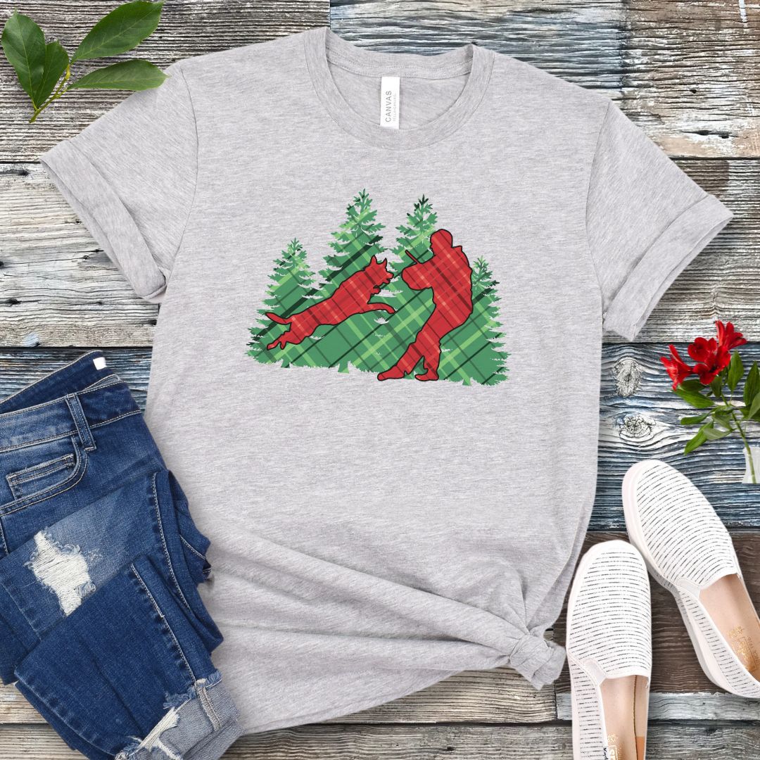 Gray t-shirt with red and green plaid holiday graphic of a Schutzhund dog and handler doing bitework, presented on a wooden surface with jeans and shoes.