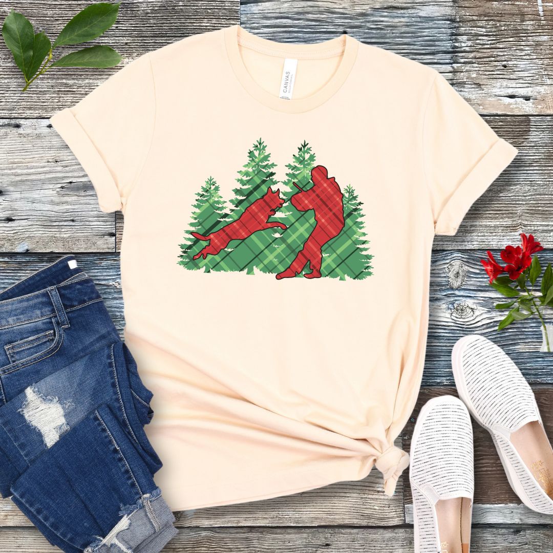 Beige t-shirt with red and green plaid holiday graphic of a Schutzhund dog and handler doing bitework, presented on a wooden surface with jeans and shoes.