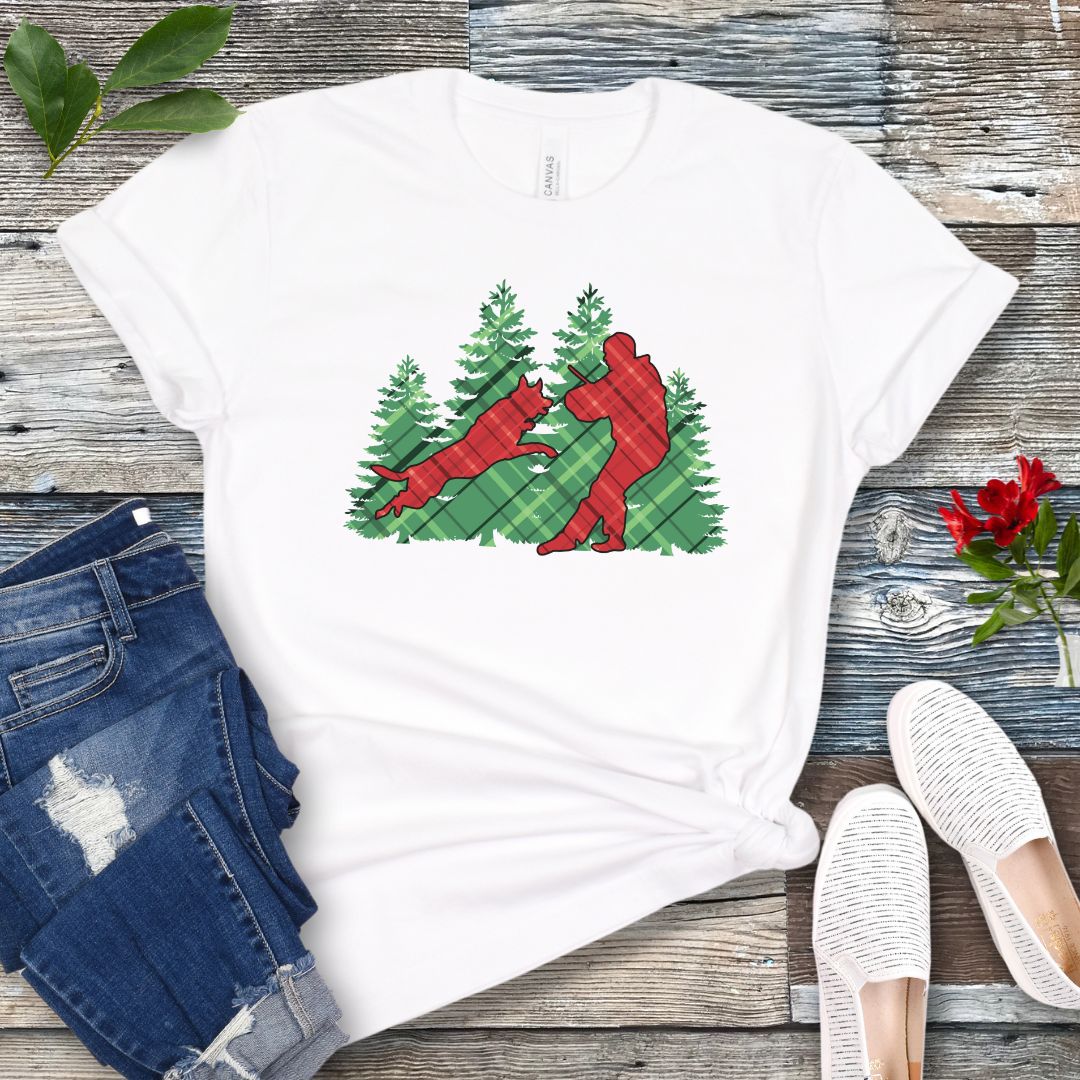 White t-shirt with red and green plaid holiday graphic of a Schutzhund dog and handler doing bitework, presented on a wooden surface with jeans and shoes.