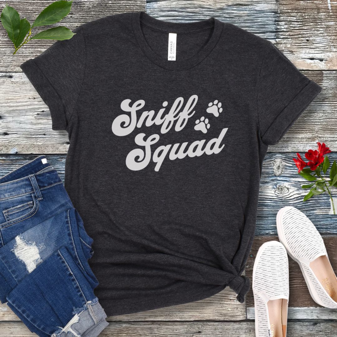 Dark grey K9 nose work t-shirt with 'Sniff Squad' text in retro font and paw prints on a wooden surface with jeans and shoes.